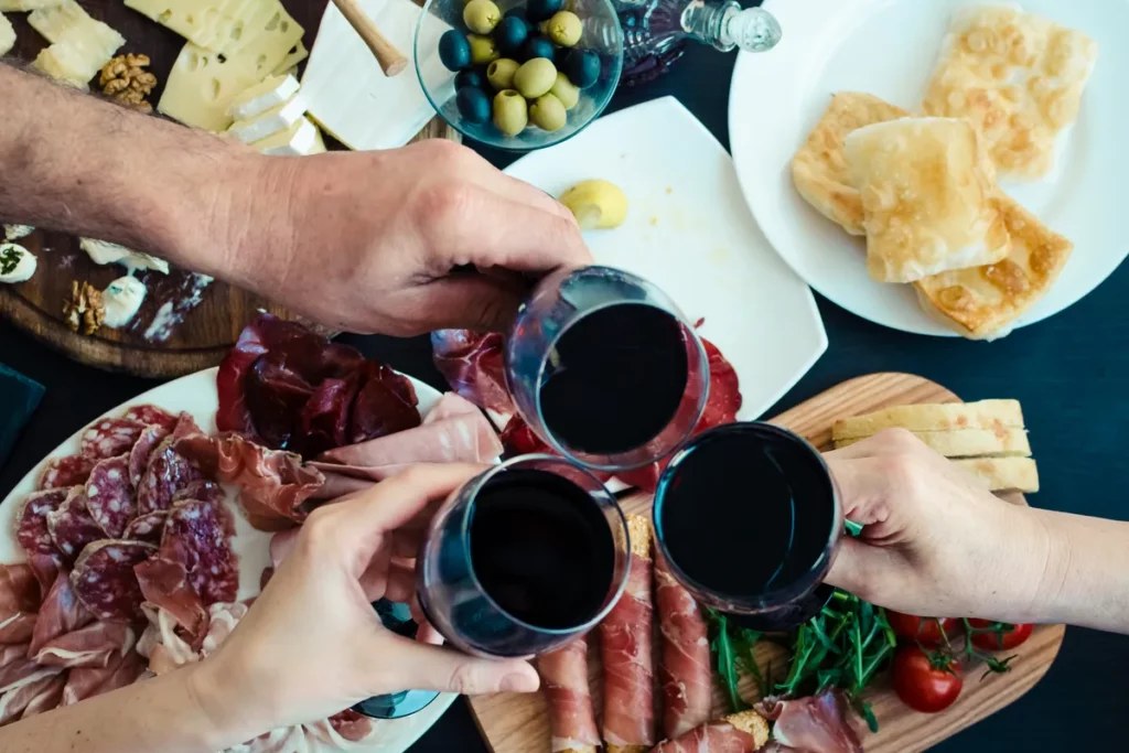 Top view of people eating and toasting with glasses of red wine. Friends or family different ages enjoying dinner concept. Delicious snack on party or picnic time. Italian style banquet.